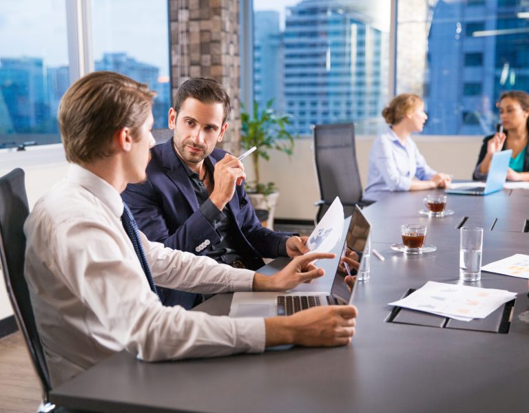Two serious young businessmen sitting in office, discussing business issues and two businesswomen working on background